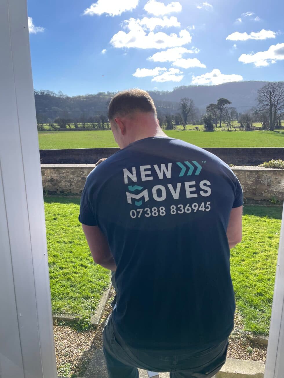 A New Moves Removals crew member wearing a branded t-shirt, seen from behind, looking out over green fields and hills from a doorway