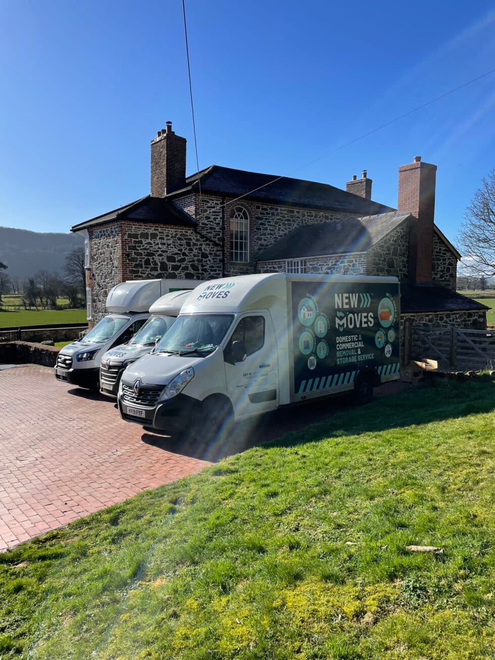 Three New Moves Removals vans parked outside a large stone house, with countryside in the background