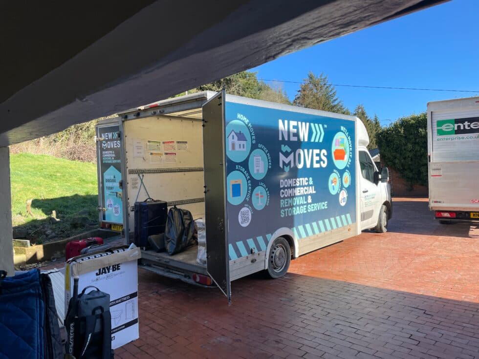 A New Moves Removals van with its side door open, parked under a carport, with luggage and boxes being loaded outside a house