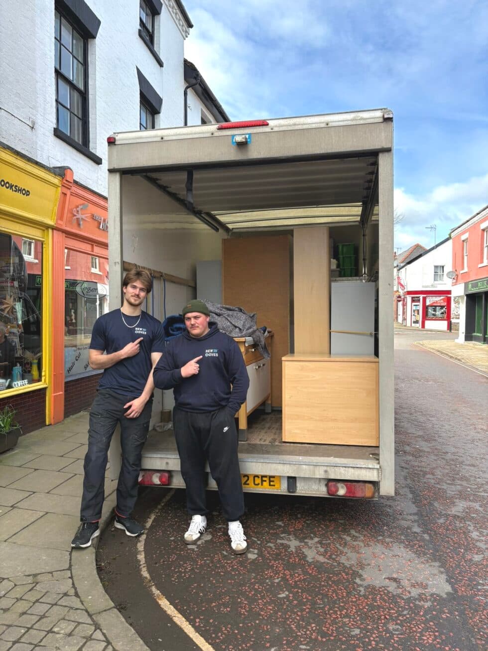 Two New Moves Removals team members standing at the back of an open removal van loaded with office furniture on a Leominster street