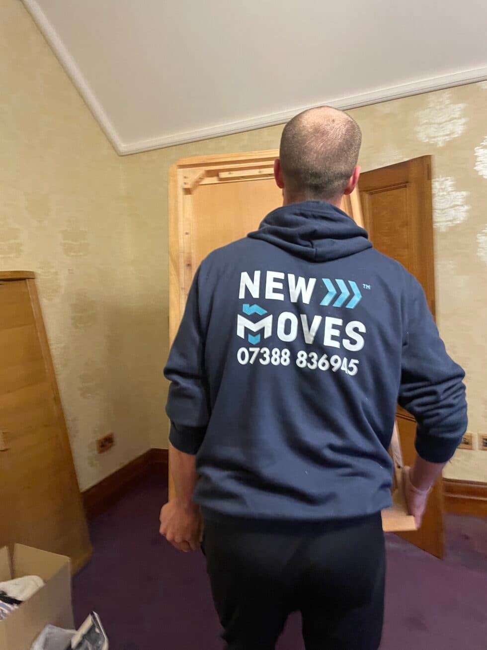 New Moves Removals team member in a branded hoodie carrying a wardrobe out of a room during a house move