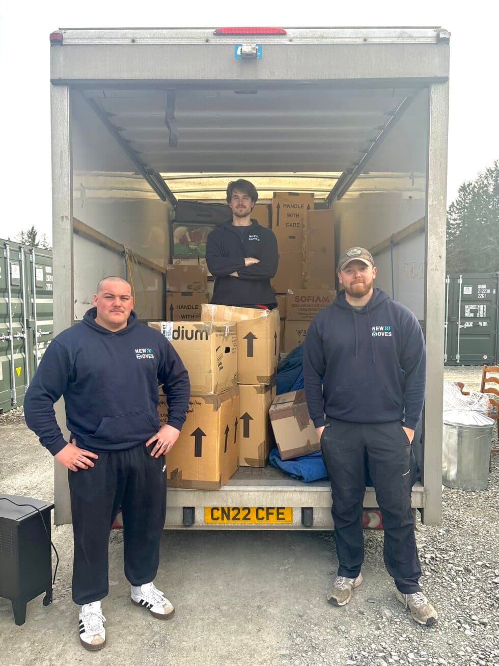 Three New Moves team members standing in front of an open removal van loaded with boxes and furniture at a storage facility