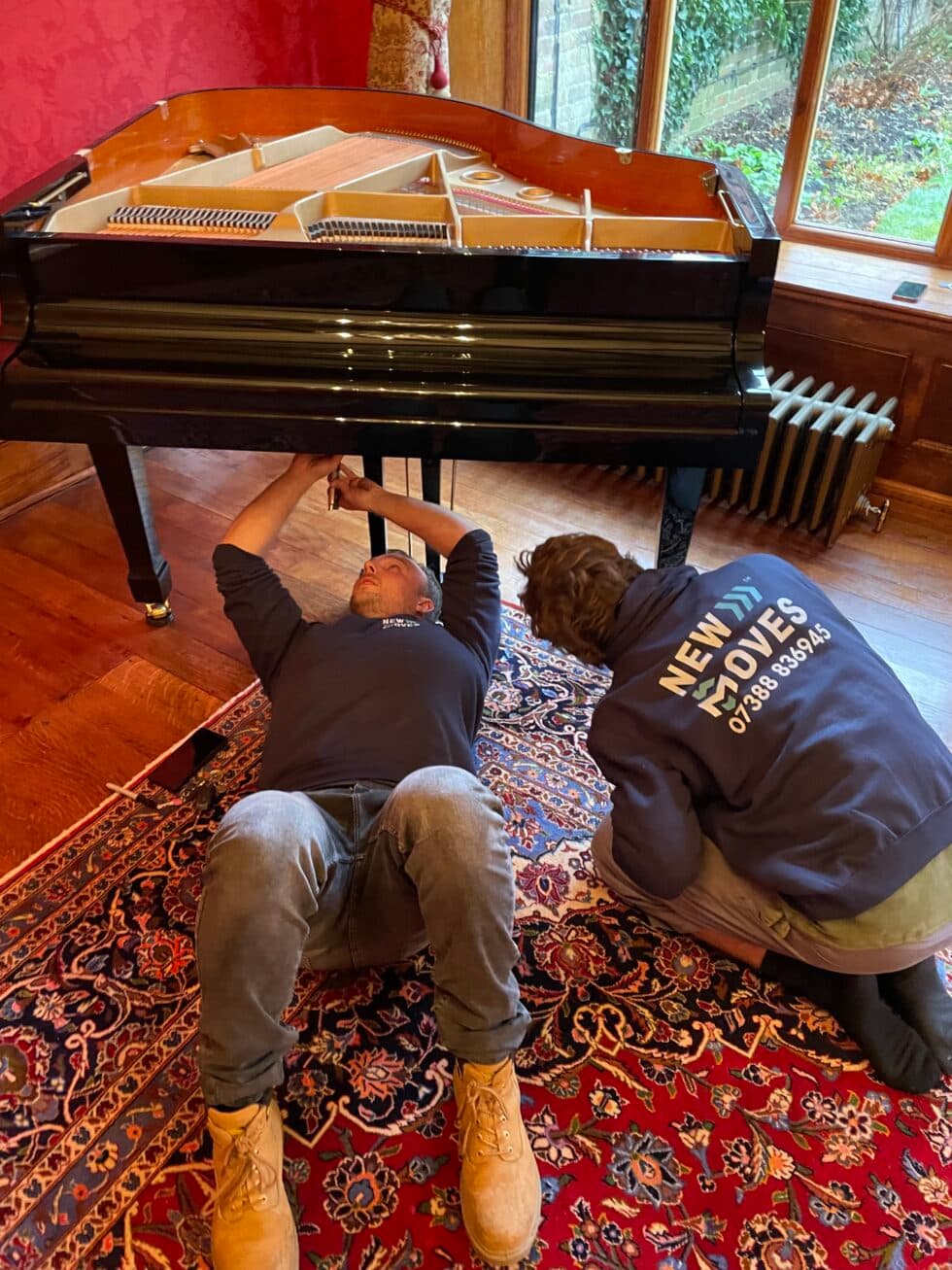 Two New Moves team members working underneath a black grand piano to prepare its legs for a move, on a Persian rug in a period room