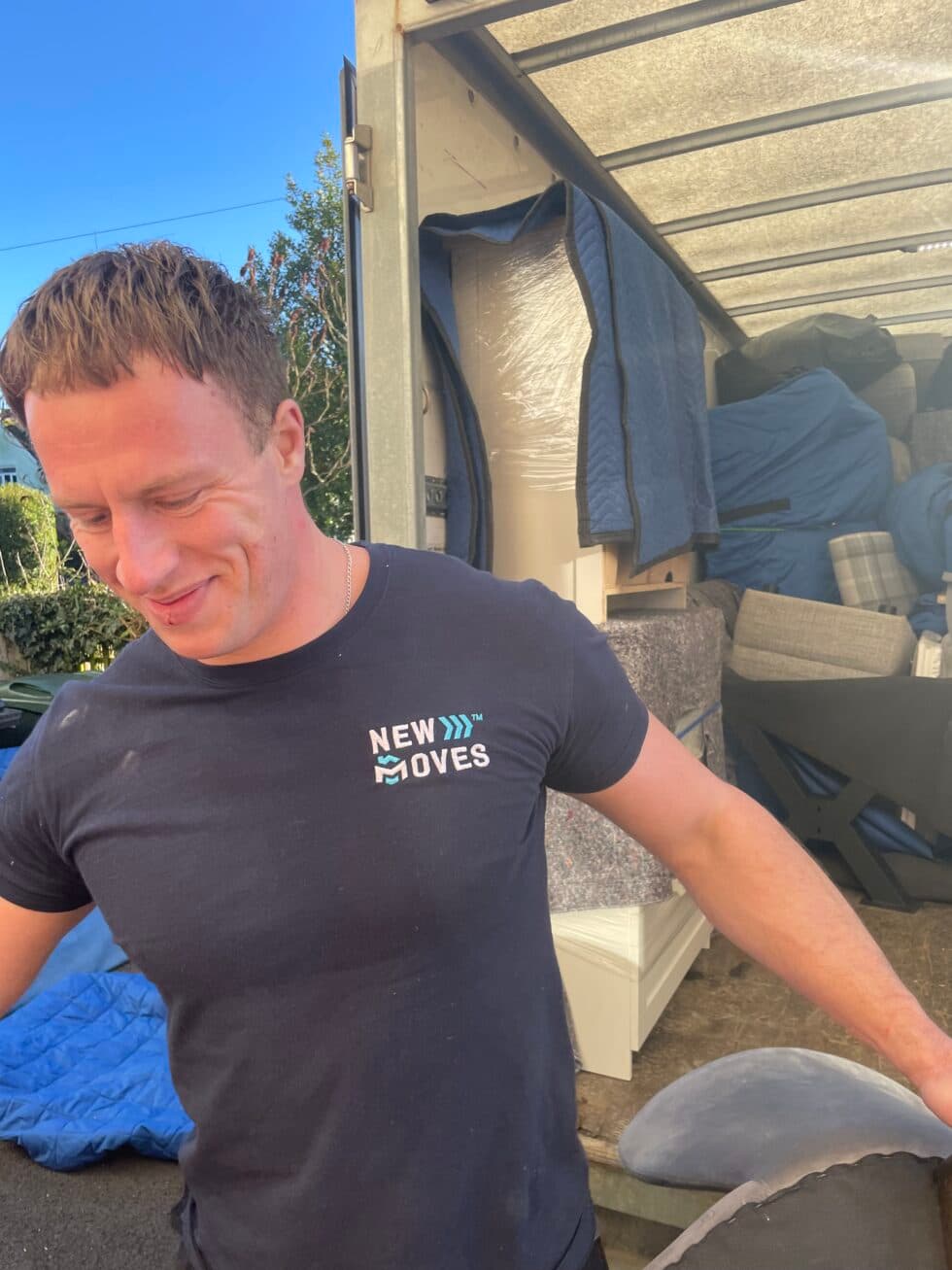 New Moves team member in a branded t-shirt in front of an open removal van packed with wrapped furniture and boxes